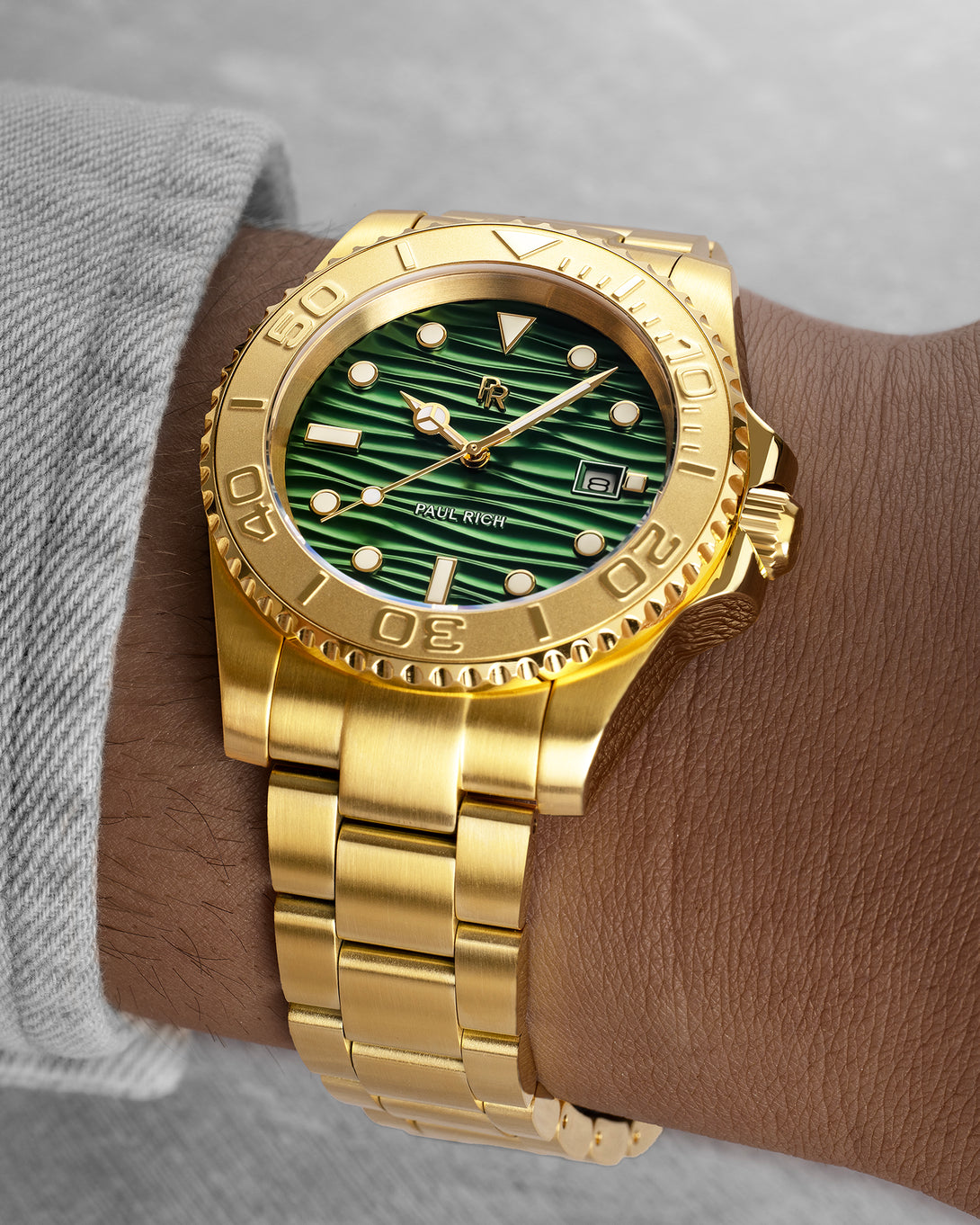 Gold-tone stainless steel watch with a green wave-textured dial shown on a wrist, highlighting the bracelet, bezel, and luminous markers. Gold-tone stainless steel watch with a green wave-textured dial shown on a wrist, highlighting the bracelet, bezel, and luminous markers.
