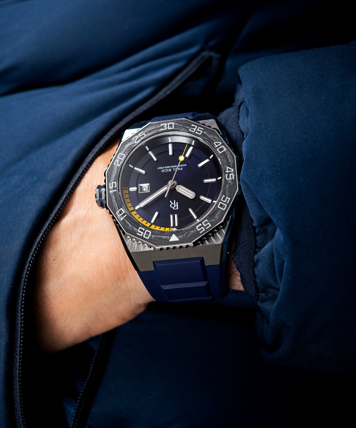 Diver-style watch with brushed steel case, black carbon bezel, blue dial, yellow depth gauge, and blue rubber strap, worn with a navy jacket. Diver-style watch with brushed steel case, black carbon bezel, blue dial, yellow depth gauge, and blue rubber strap, worn with a navy jacket.