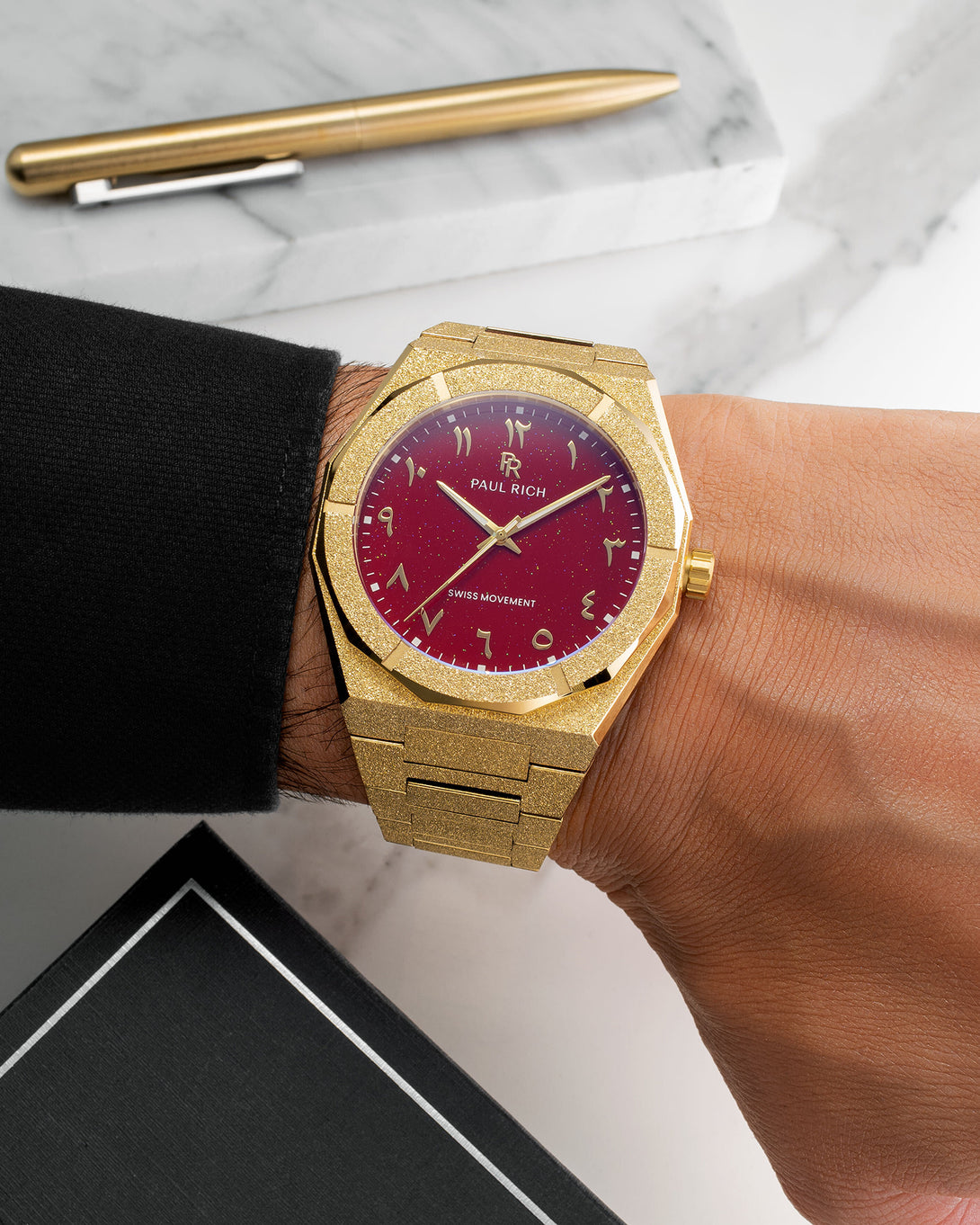 Paul Rich watch with frosted gold case and red aventurine dial with Arabic numerals, worn with a black cuff on a marble desk. Paul Rich watch with frosted gold case and red aventurine dial with Arabic numerals, worn with a black cuff on a marble desk.