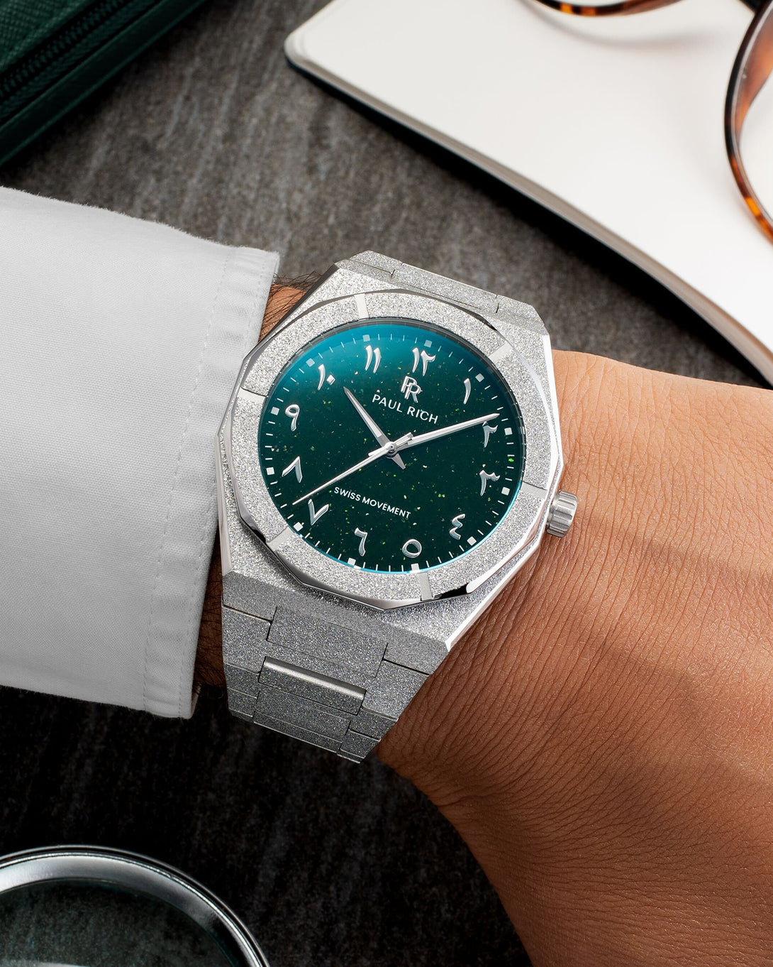 Paul Rich watch with frosted silver case and green aventurine dial with Arabic numerals, worn with a white cuff on a dark tabletop. Paul Rich watch with frosted silver case and green aventurine dial with Arabic numerals, worn with a white cuff on a dark tabletop.