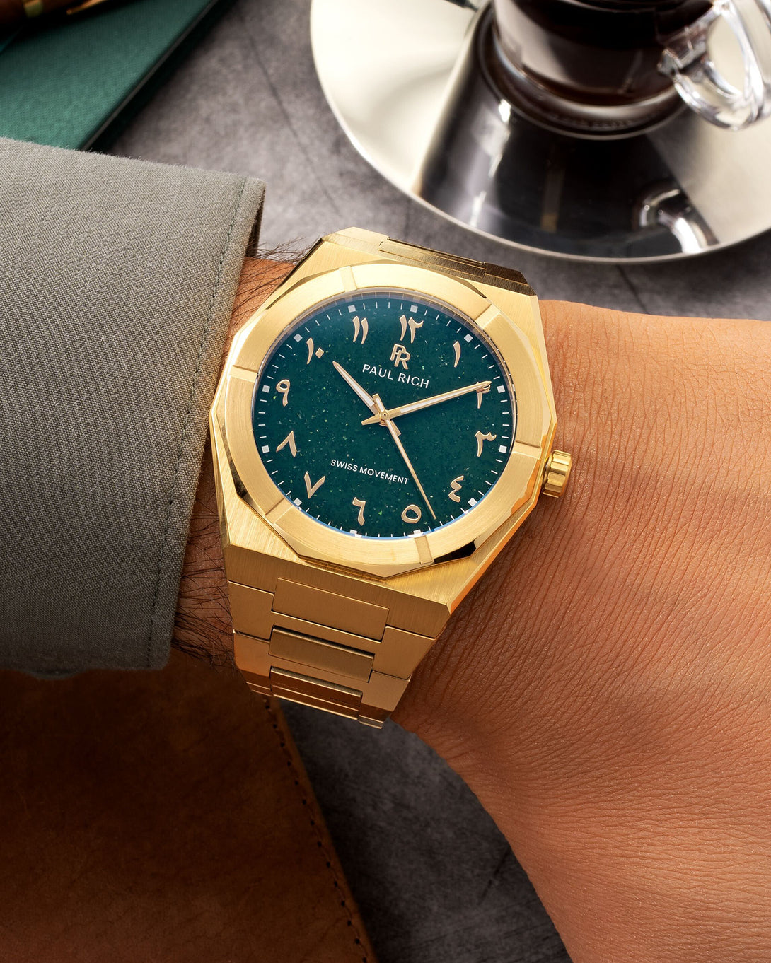 Paul Rich watch with brushed gold case and green dial featuring Arabic numerals, worn with a sage cuff beside a cup of espresso. Paul Rich watch with brushed gold case and green dial featuring Arabic numerals, worn with a sage cuff beside a cup of espresso.