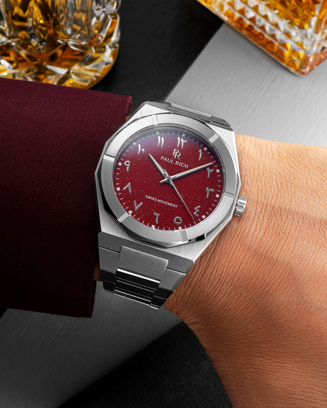Paul Rich watch with brushed silver case and red dial with Arabic numerals, worn with a burgundy sleeve near crystal whiskey glasses. Paul Rich watch with brushed silver case and red dial with Arabic numerals, worn with a burgundy sleeve near crystal whiskey glasses.