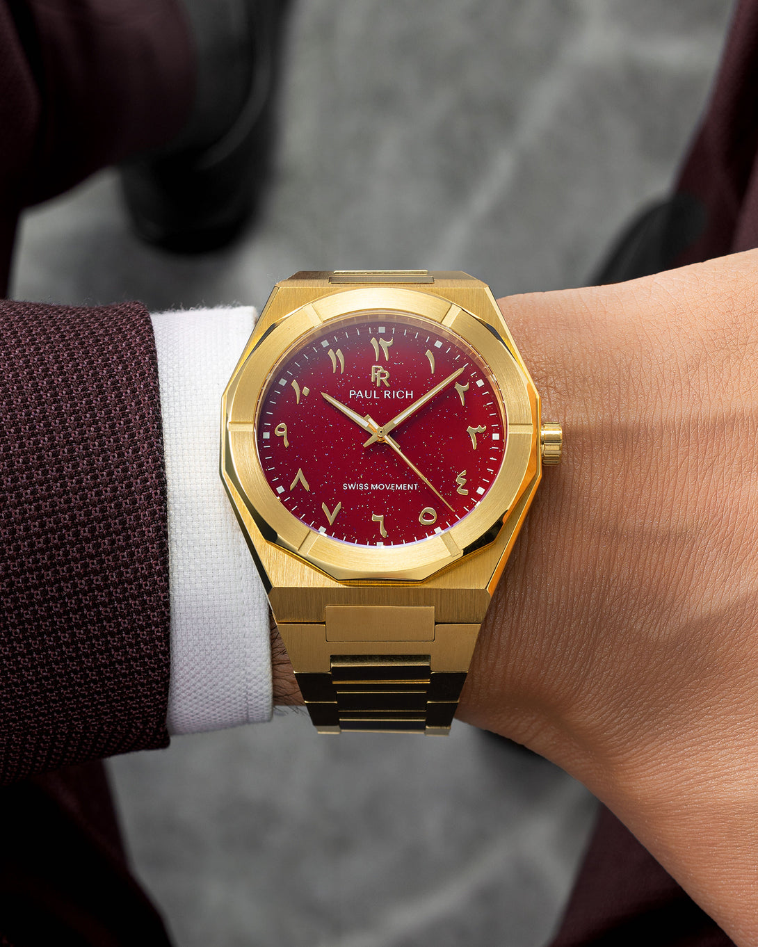 Paul Rich watch with brushed gold case and red aventurine dial featuring Arabic numerals, worn with a burgundy textured blazer. Paul Rich watch with brushed gold case and red aventurine dial featuring Arabic numerals, worn with a burgundy textured blazer.