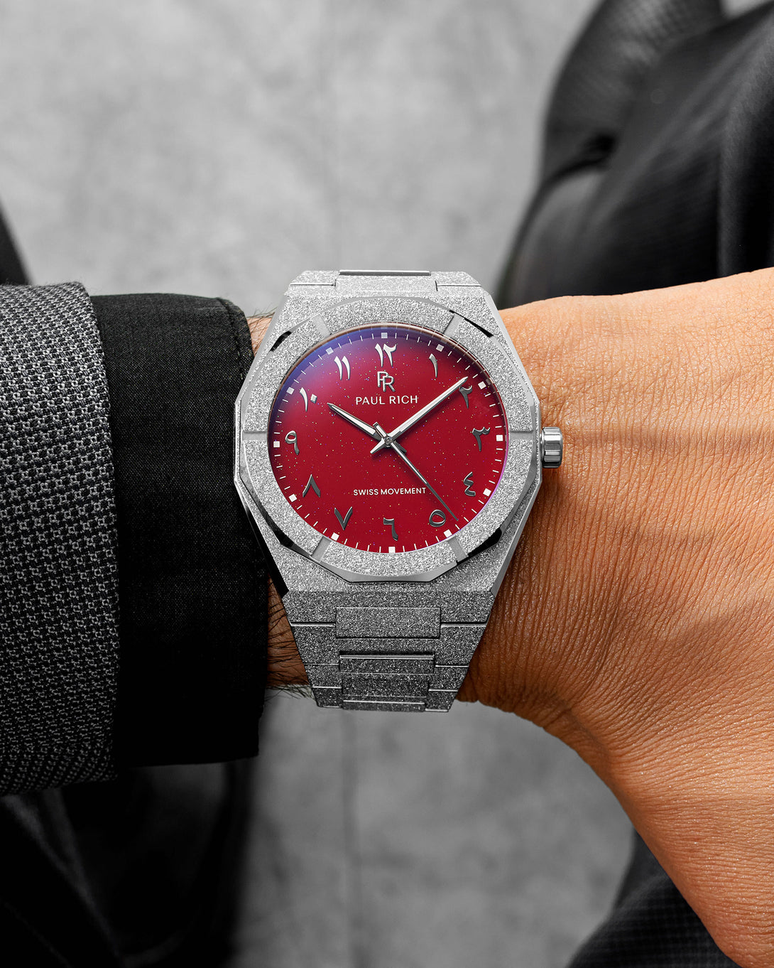 Paul Rich watch with frosted silver case and red aventurine dial with Arabic numerals, worn with a black and gray textured suit. Paul Rich watch with frosted silver case and red aventurine dial with Arabic numerals, worn with a black and gray textured suit.