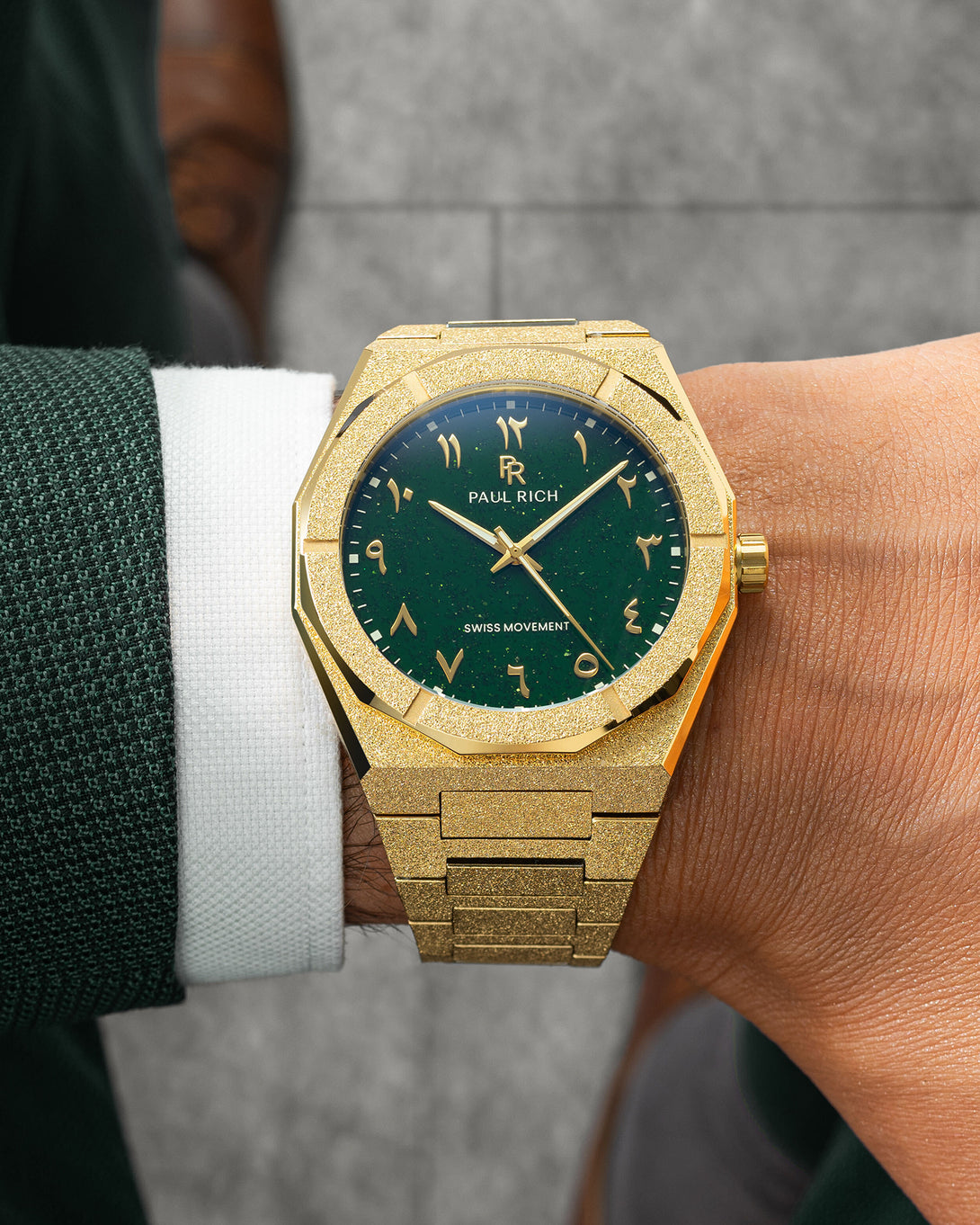 Paul Rich watch with frosted gold case and green aventurine dial with Arabic numerals, worn with a dark green textured suit and white cuff. Paul Rich watch with frosted gold case and green aventurine dial with Arabic numerals, worn with a dark green textured suit and white cuff.