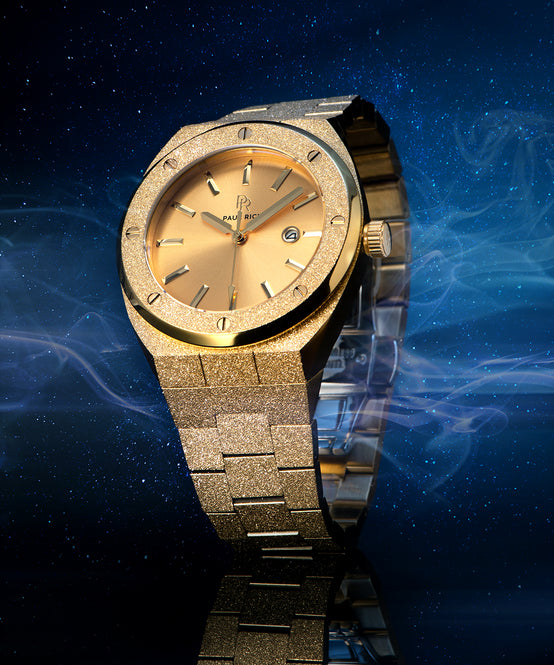 Paul Rich watch with frosted gold case and bracelet, metallic champagne gold sunray dial, and date window, displayed against a starry night sky with mist. Paul Rich watch with frosted gold case and bracelet, metallic champagne gold sunray dial, and date window, displayed against a starry night sky with mist.