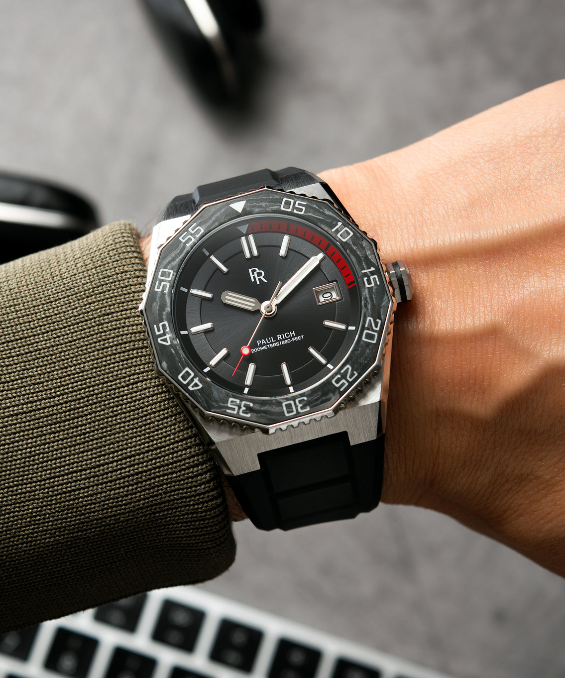 Paul Rich dive watch with black sunray dial and red dive timer worn on wrist, paired with green cuff and visible laptop keyboard. Paul Rich dive watch with black sunray dial and red dive timer worn on wrist, paired with green cuff and visible laptop keyboard.
