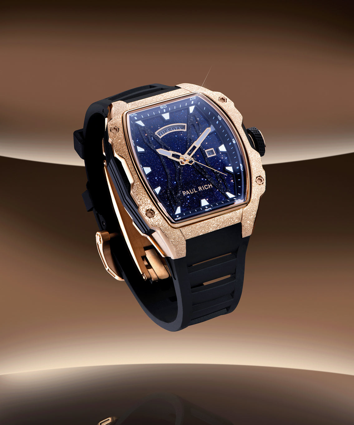 Paul Rich watch with frosted rose gold tonneau case, blue aventurine dial, day-date display, and black strap, floating over a bronze gradient background. Paul Rich watch with frosted rose gold tonneau case, blue aventurine dial, day-date display, and black strap, floating over a bronze gradient background.