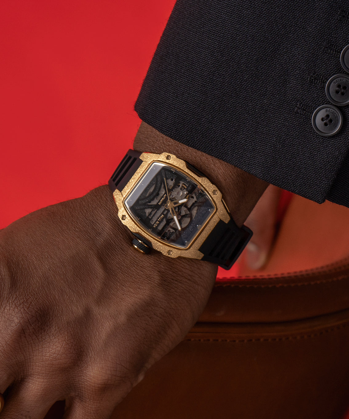 Man wearing a Paul Rich watch with frosted gold tonneau case and black skeleton dial, paired with black suit, against a red backdrop. Man wearing a Paul Rich watch with frosted gold tonneau case and black skeleton dial, paired with black suit, against a red backdrop.