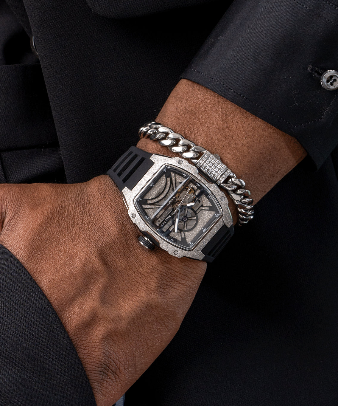 Man wearing a Paul Rich watch with frosted silver tonneau case and skeleton dial, paired with silver Cuban link bracelet over black suit. Man wearing a Paul Rich watch with frosted silver tonneau case and skeleton dial, paired with silver Cuban link bracelet over black suit.