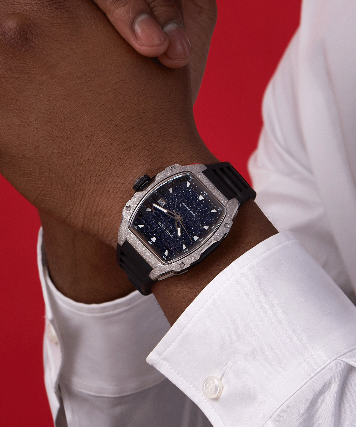 Paul Rich watch with tonneau case, frosted silver bezel, and blue aventurine dial, worn on wrist with black strap and white cuff, against red background. Paul Rich watch with tonneau case, frosted silver bezel, and blue aventurine dial, worn on wrist with black strap and white cuff, against red background.