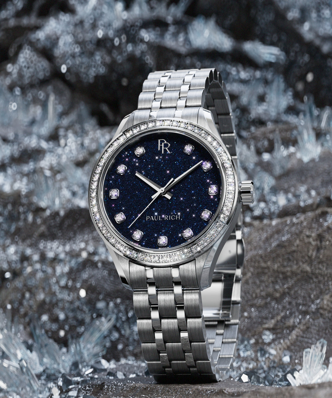 Silver-tone Paul Rich watch with deep blue aventurine dial, diamond hour markers, and gem-set bezel, displayed upright on sparkling crystal rocks. Silver-tone Paul Rich watch with deep blue aventurine dial, diamond hour markers, and gem-set bezel, displayed upright on sparkling crystal rocks.