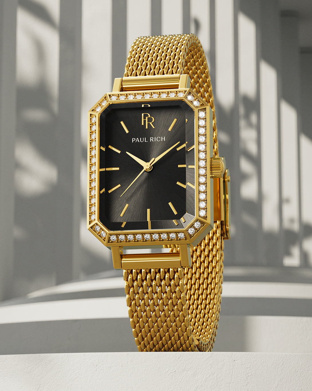 Gold-tone Paul Rich watch with black sunray dial, gem-set octagonal bezel, and gold mesh strap, displayed upright on a white surface with vertical shadow patterns in the background. Gold-tone Paul Rich watch with black sunray dial, gem-set octagonal bezel, and gold mesh strap, displayed upright on a white surface with vertical shadow patterns in the background.