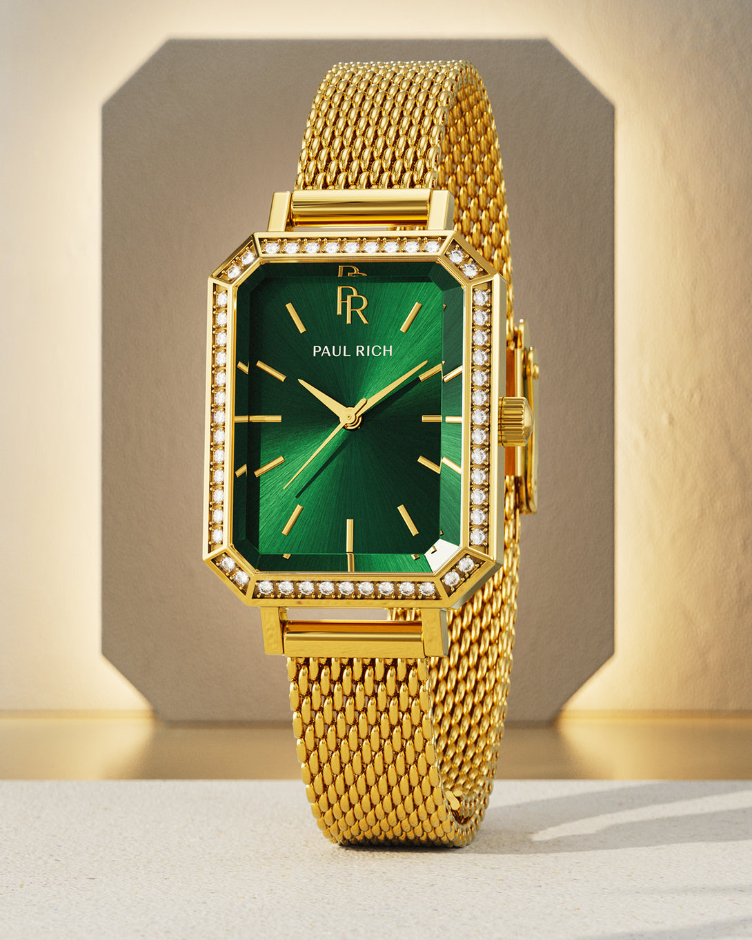 Gold-tone Paul Rich watch with emerald green sunray dial, gem-set octagonal bezel, and gold mesh strap, displayed upright against a beige geometric backdrop with soft shadows. Gold-tone Paul Rich watch with emerald green sunray dial, gem-set octagonal bezel, and gold mesh strap, displayed upright against a beige geometric backdrop with soft shadows.