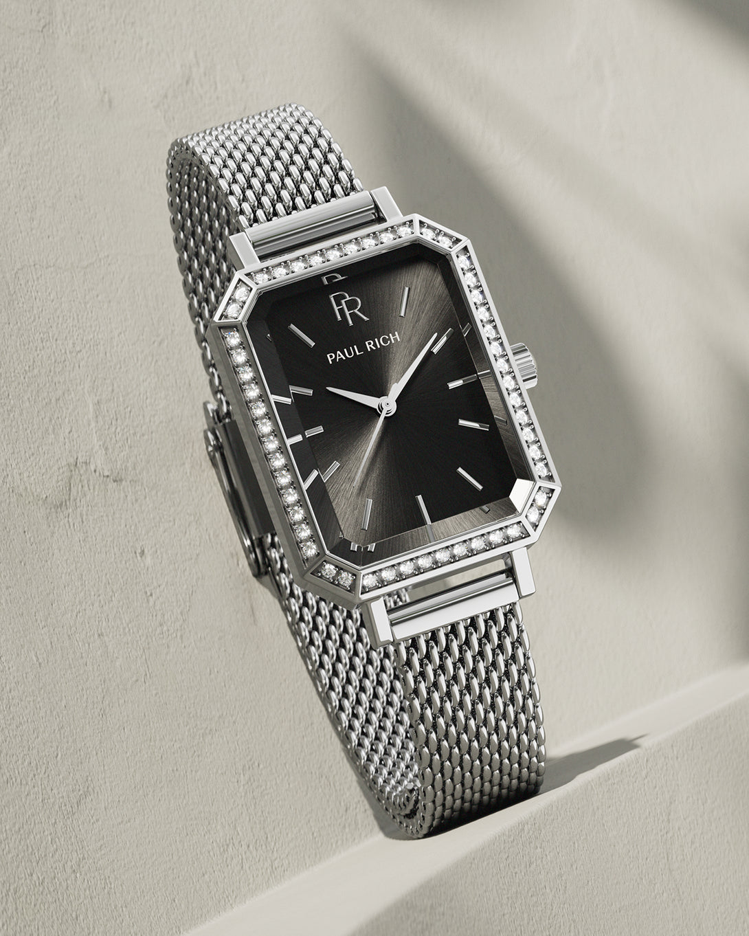 Silver-tone Paul Rich watch with black sunray dial, gem-set octagonal bezel, and silver mesh strap, displayed leaning against a textured beige wall with soft shadowing. Silver-tone Paul Rich watch with black sunray dial, gem-set octagonal bezel, and silver mesh strap, displayed leaning against a textured beige wall with soft shadowing.