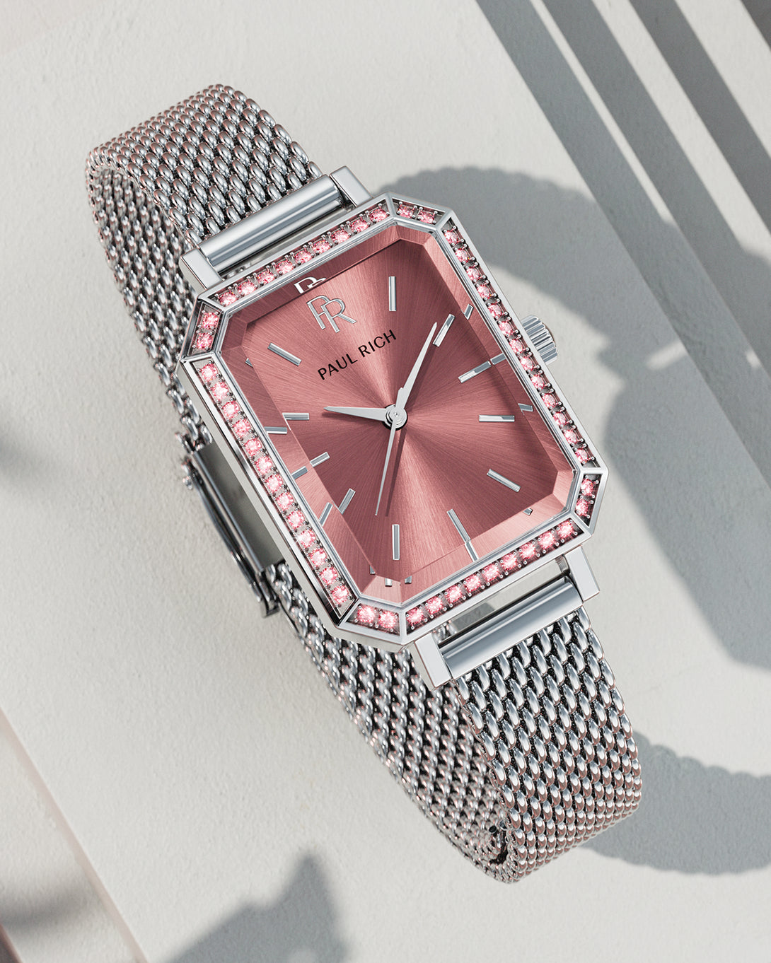 Silver-tone Paul Rich watch with rose pink sunray dial, pink gem-set octagonal bezel, and silver mesh strap, displayed on a textured off-white surface with sharp shadows. Silver-tone Paul Rich watch with rose pink sunray dial, pink gem-set octagonal bezel, and silver mesh strap, displayed on a textured off-white surface with sharp shadows.