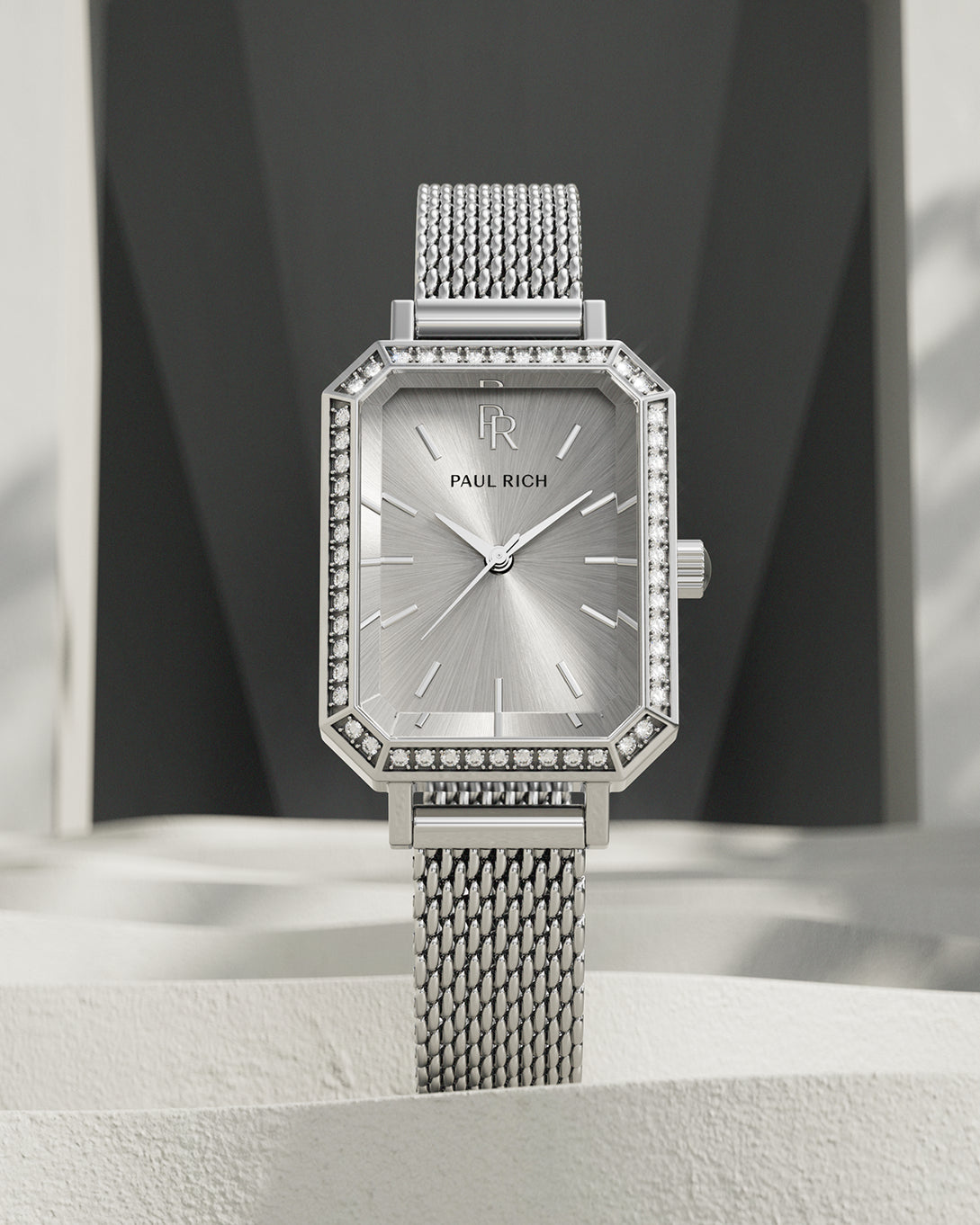 Silver-tone Paul Rich watch with silver sunray dial, gem-set octagonal bezel, and silver mesh strap, displayed upright on a textured off-white surface with geometric dark gray and white background. Silver-tone Paul Rich watch with silver sunray dial, gem-set octagonal bezel, and silver mesh strap, displayed upright on a textured off-white surface with geometric dark gray and white background.