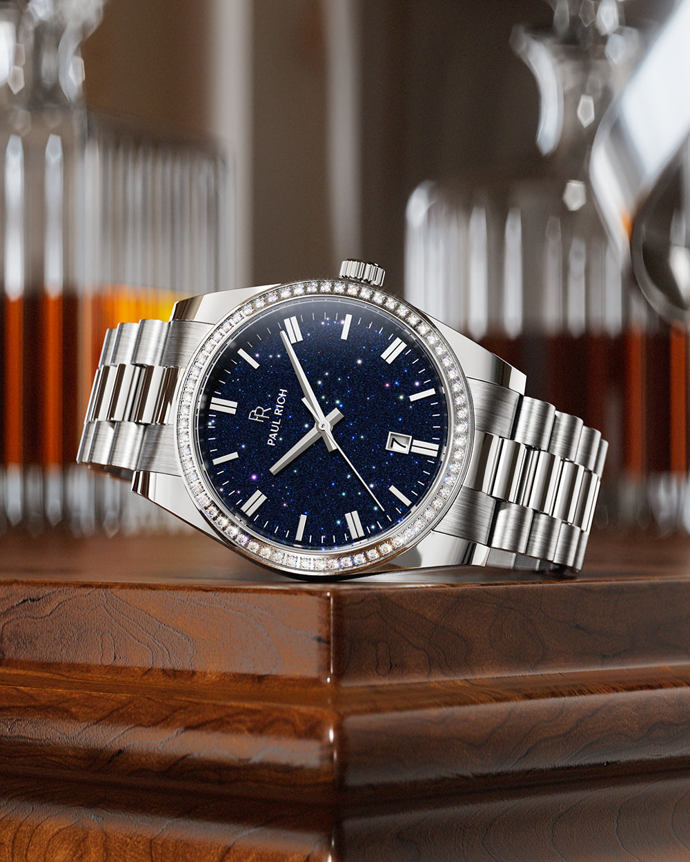 Silver wristwatch with a blue speckled dial and crystal-set bezel resting on a polished wooden surface, with glass decanters in the background. Silver wristwatch with a blue speckled dial and crystal-set bezel resting on a polished wooden surface, with glass decanters in the background.