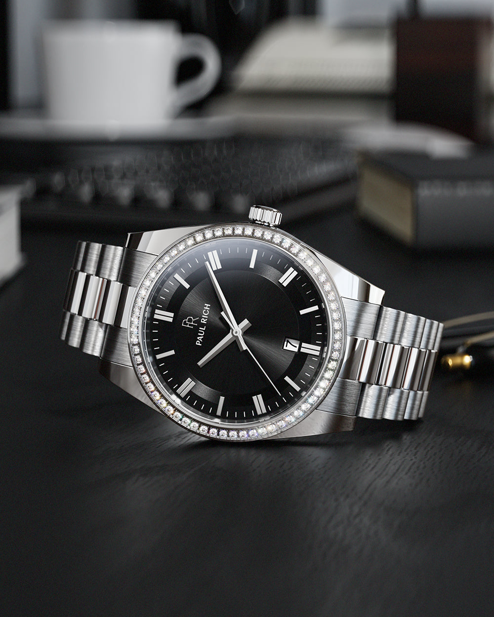Silver-tone Paul Rich watch with black sunray dial, diamond-set bezel, and silver link bracelet, displayed on a black desk surface with a white coffee cup, keyboard, and books blurred in the background. Silver-tone Paul Rich watch with black sunray dial, diamond-set bezel, and silver link bracelet, displayed on a black desk surface with a white coffee cup, keyboard, and books blurred in the background.