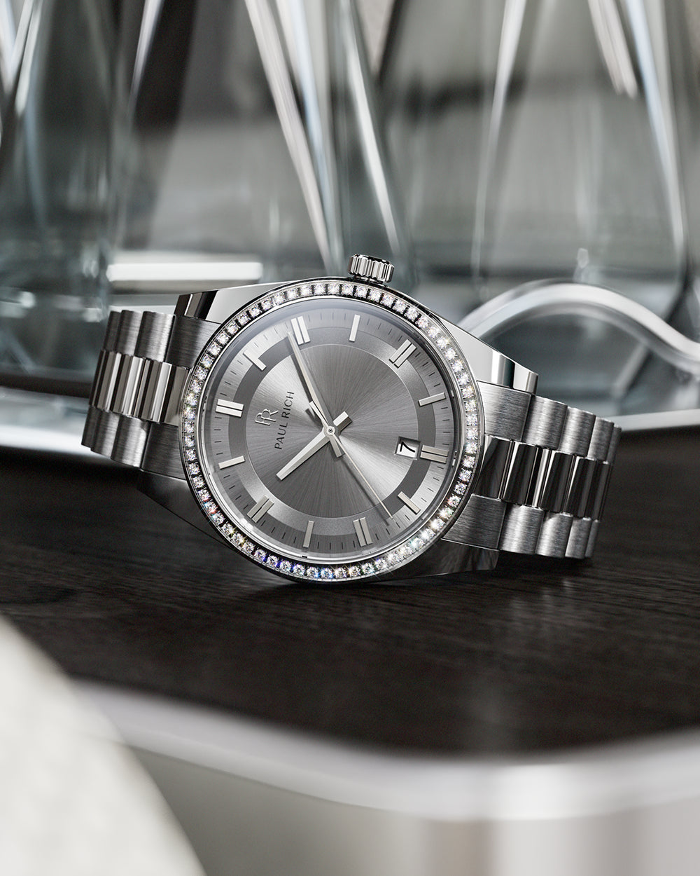 Silver-tone Paul Rich watch with silver sunray dial, diamond-set bezel, and silver link bracelet, displayed on a dark wooden surface with reflective glass decanters in the background. Silver-tone Paul Rich watch with silver sunray dial, diamond-set bezel, and silver link bracelet, displayed on a dark wooden surface with reflective glass decanters in the background.