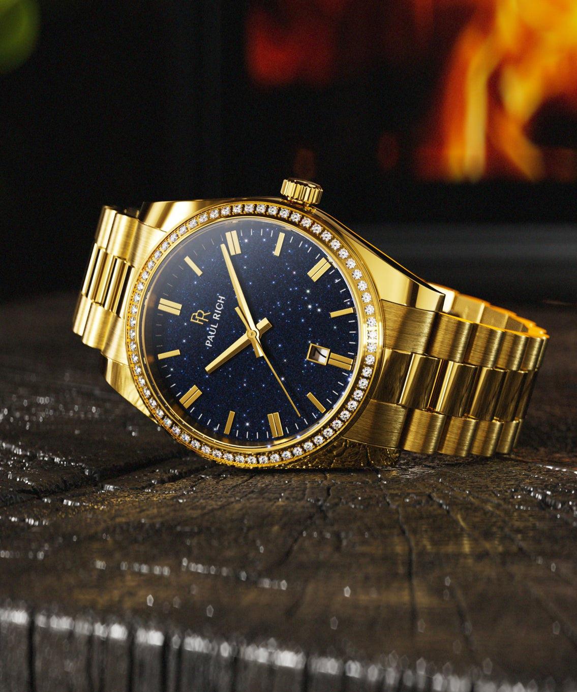 Gold Paul Rich watch with a blue aventurine dial and gem-set bezel, resting on wood with a fireplace glowing in the background. Gold Paul Rich watch with a blue aventurine dial and gem-set bezel, resting on wood with a fireplace glowing in the background.