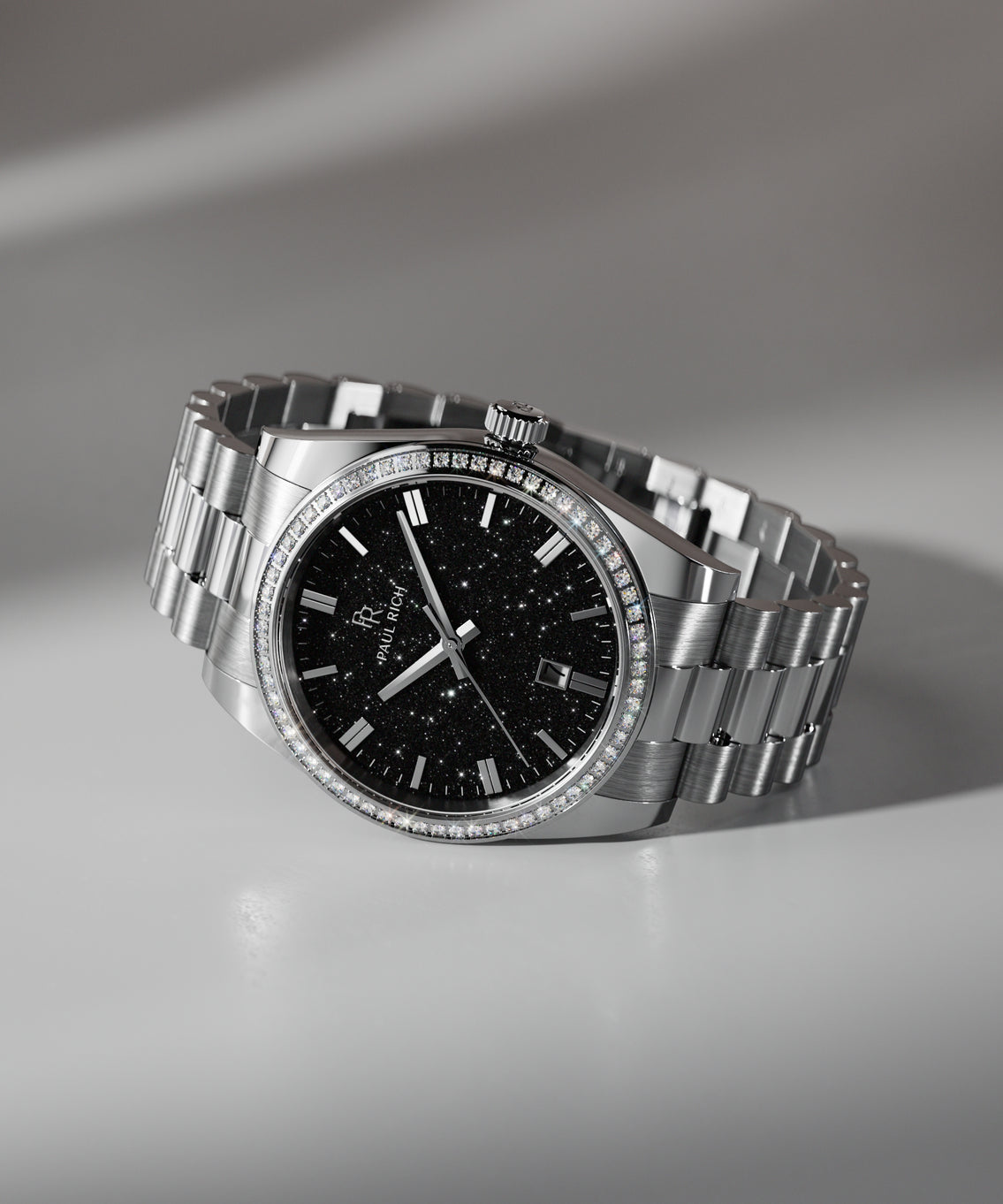 Silver wristwatch with a black aventurine dial, crystal-set bezel, and metal bracelet displayed on a white surface with soft shadowing. Silver wristwatch with a black aventurine dial, crystal-set bezel, and metal bracelet displayed on a white surface with soft shadowing.
