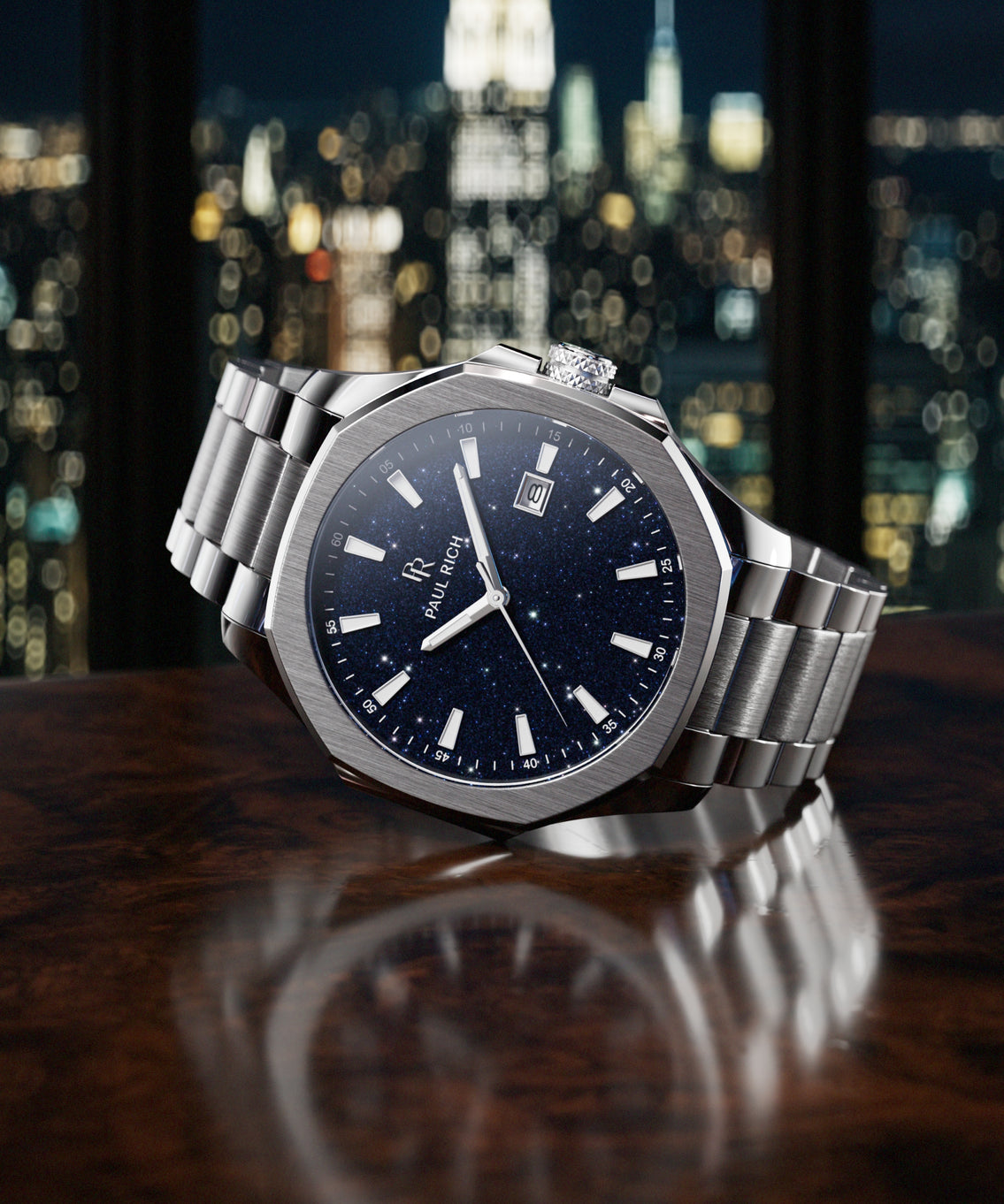 Paul Rich watch with brushed silver case and blue aventurine dial resting on dark wood surface, with NYC skyline at night in background. Paul Rich watch with brushed silver case and blue aventurine dial resting on dark wood surface, with NYC skyline at night in background.