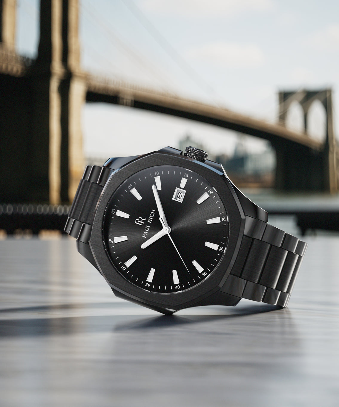 Black Paul Rich watch with a sunray dial and steel bracelet, styled on a table with the Brooklyn Bridge in the background. Black Paul Rich watch with a sunray dial and steel bracelet, styled on a table with the Brooklyn Bridge in the background.