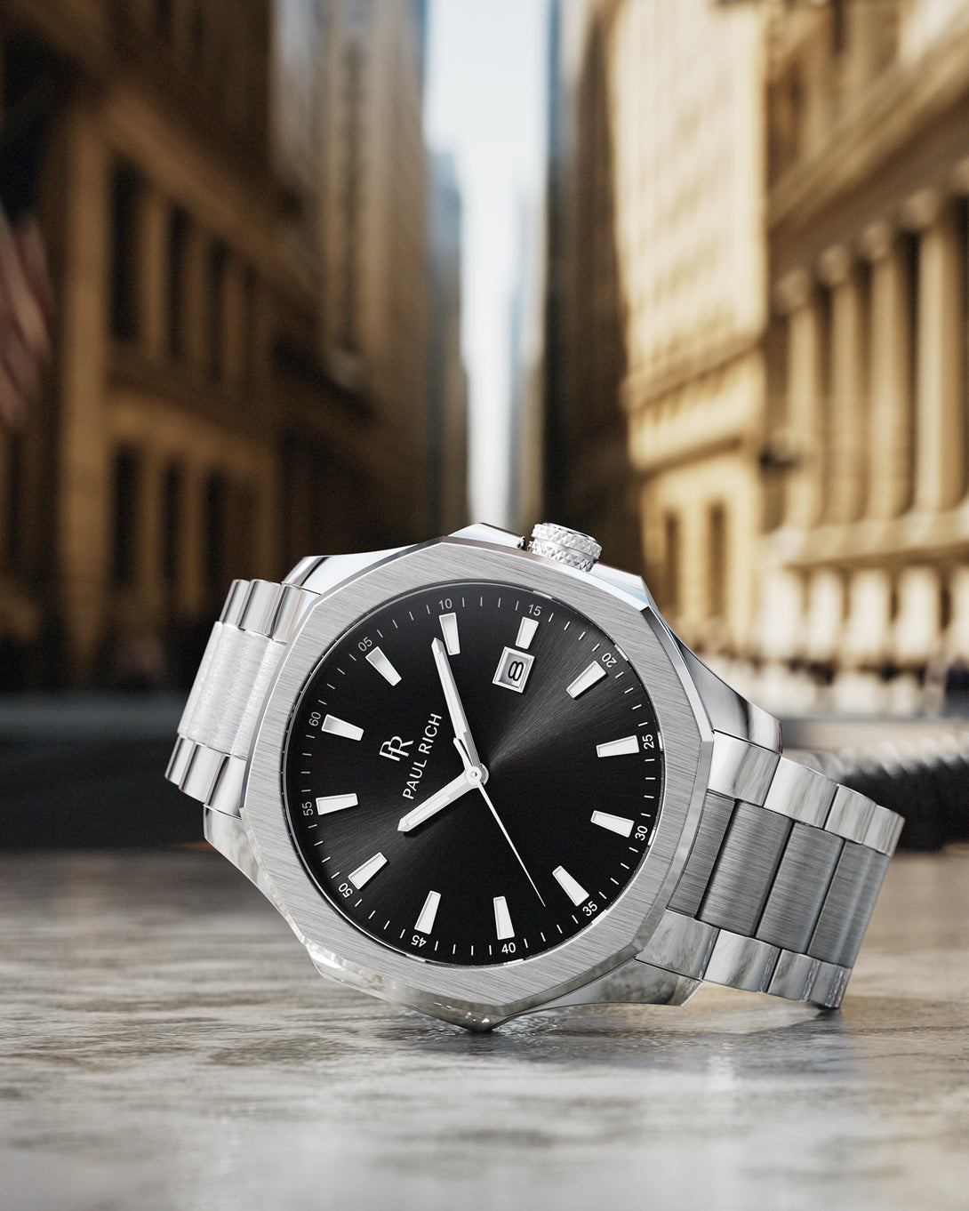 Paul Rich silver watch with black sunray dial and steel bracelet, photographed close-up on Wall Street with skyscrapers in background. Paul Rich silver watch with black sunray dial and steel bracelet, photographed close-up on Wall Street with skyscrapers in background.