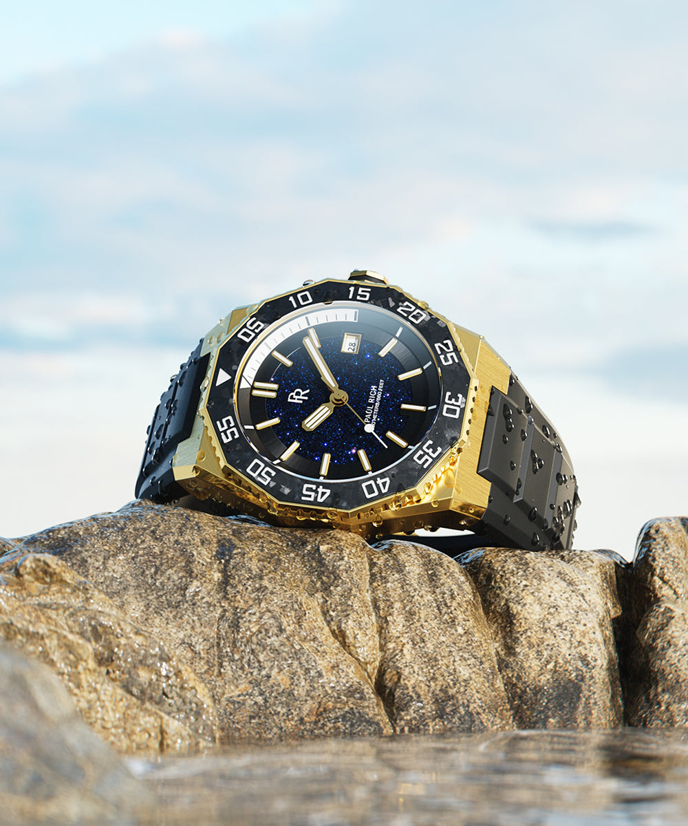 Paul Rich dive watch with gold case and aventurine dial resting on wet rock by the sea, droplets on case, with sky in background. Paul Rich dive watch with gold case and aventurine dial resting on wet rock by the sea, droplets on case, with sky in background.