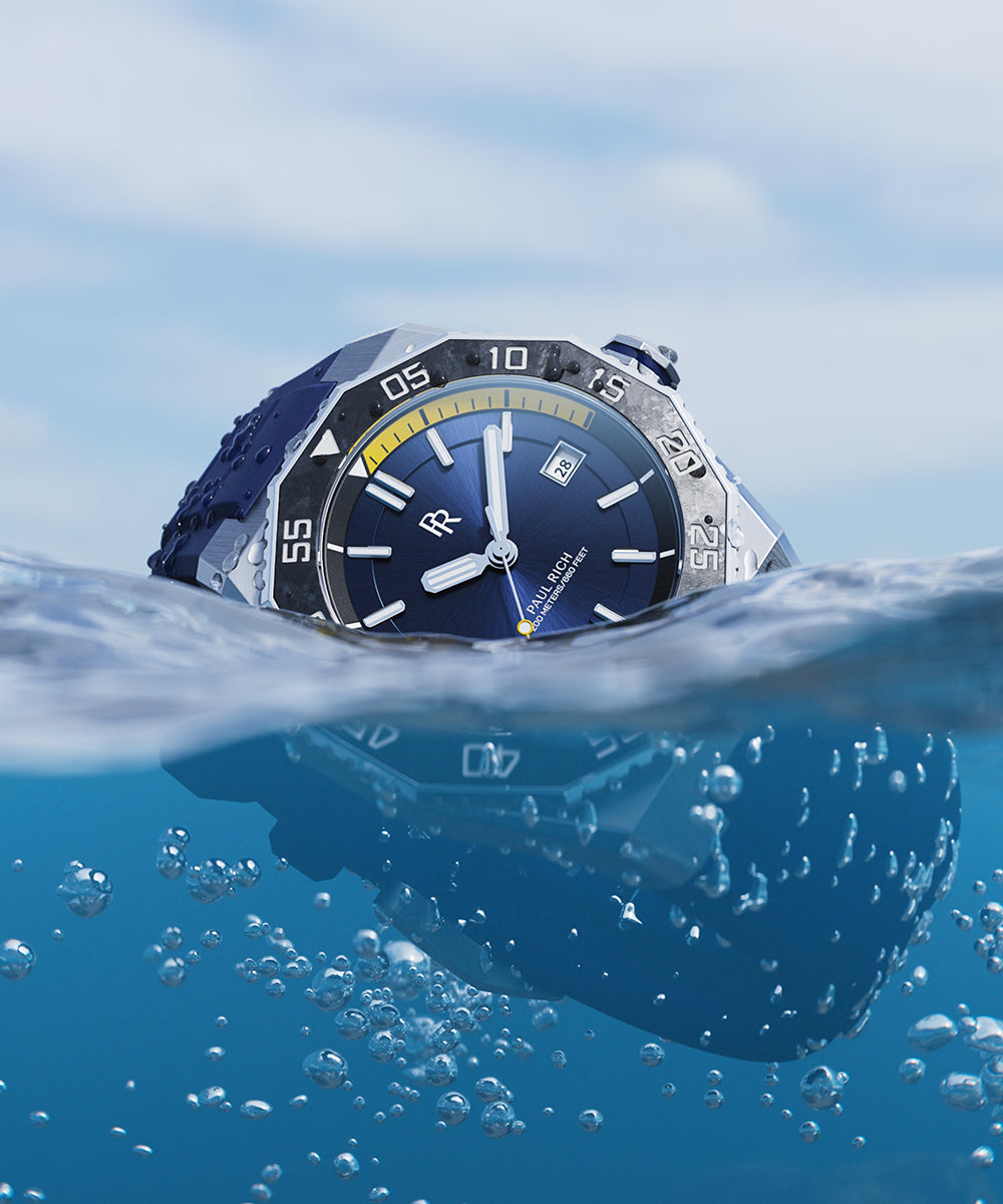Close-up of a Paul Rich dive watch half-submerged in water, blue dial and bezel visible above surface, bubbles rising below. Close-up of a Paul Rich dive watch half-submerged in water, blue dial and bezel visible above surface, bubbles rising below.