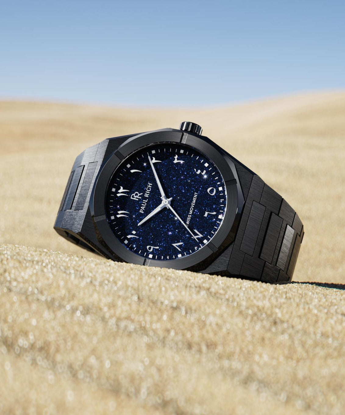 Paul Rich watch with brushed black case and blue aventurine dial with Arabic numerals, displayed on golden sand under a clear sky. Paul Rich watch with brushed black case and blue aventurine dial with Arabic numerals, displayed on golden sand under a clear sky.