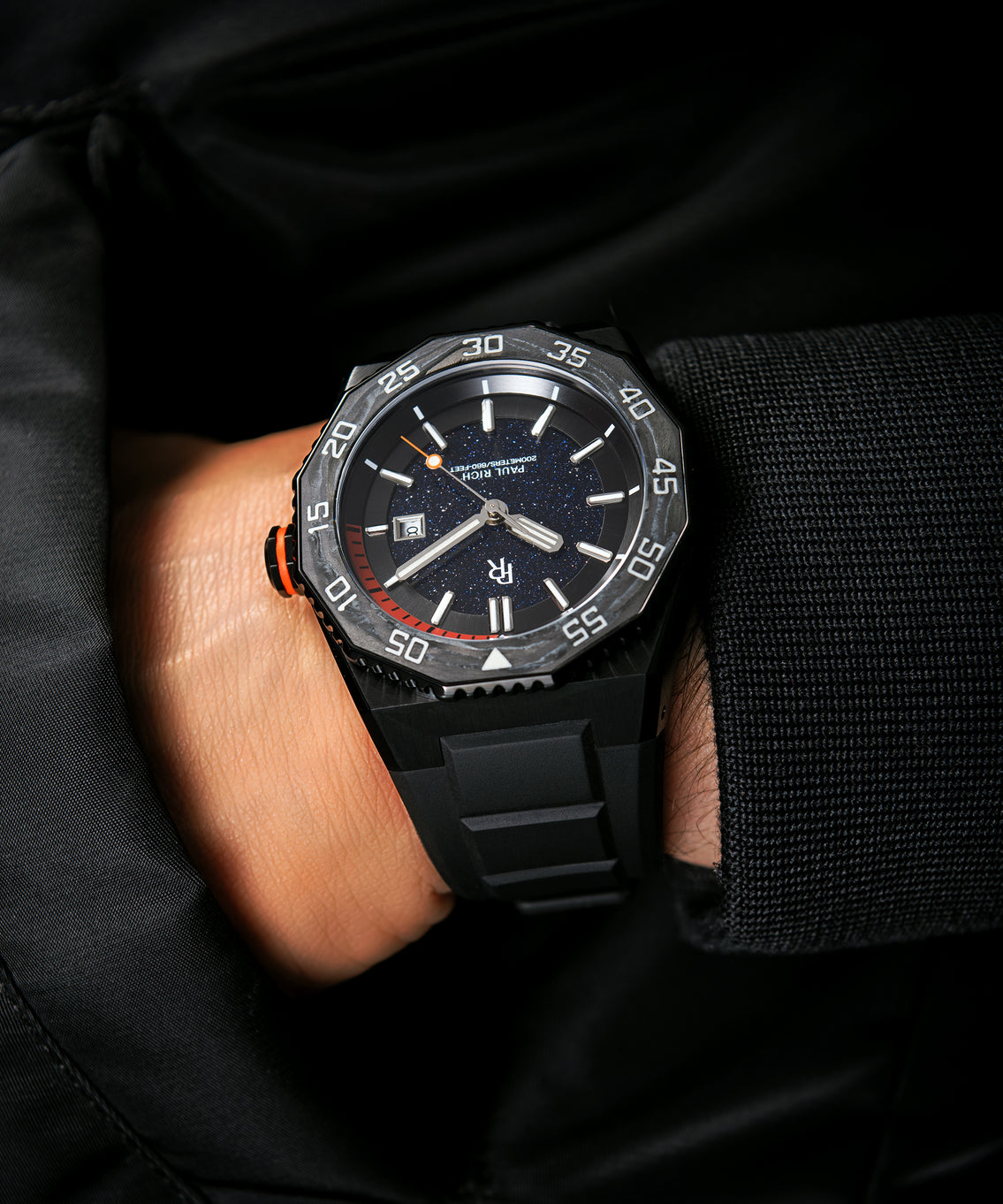 Paul Rich dive watch with aventurine dial and orange dive timer worn on wrist, paired with a black textured jacket sleeve. Paul Rich dive watch with aventurine dial and orange dive timer worn on wrist, paired with a black textured jacket sleeve.