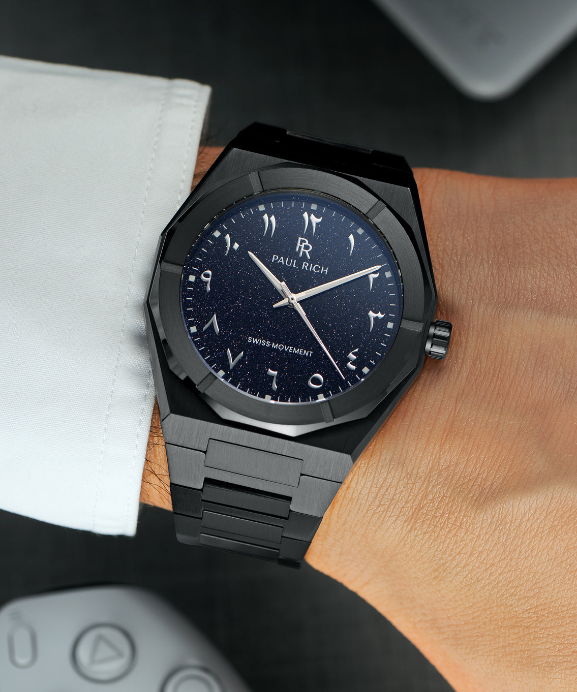 Paul Rich watch with brushed black case and blue aventurine dial with Arabic numerals, worn with a white cuff over a dark surface. Paul Rich watch with brushed black case and blue aventurine dial with Arabic numerals, worn with a white cuff over a dark surface.