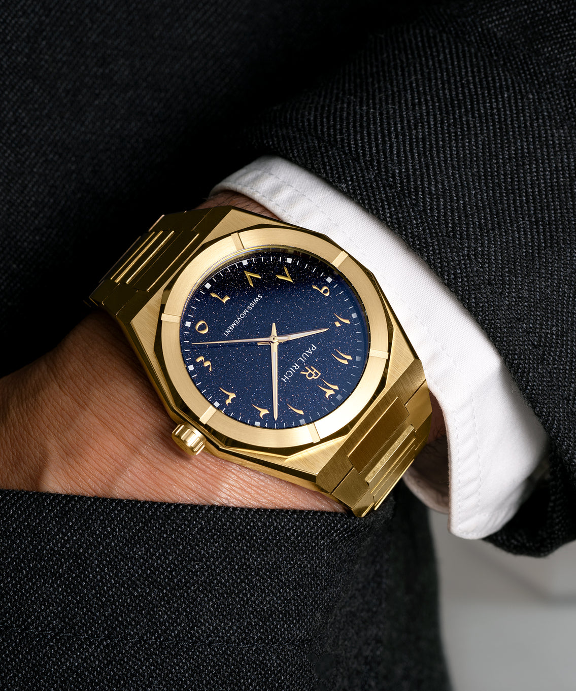 Paul Rich watch with brushed gold case and blue aventurine dial with Arabic numerals, worn with a black suit and white cuff, hand in pocket. Paul Rich watch with brushed gold case and blue aventurine dial with Arabic numerals, worn with a black suit and white cuff, hand in pocket.