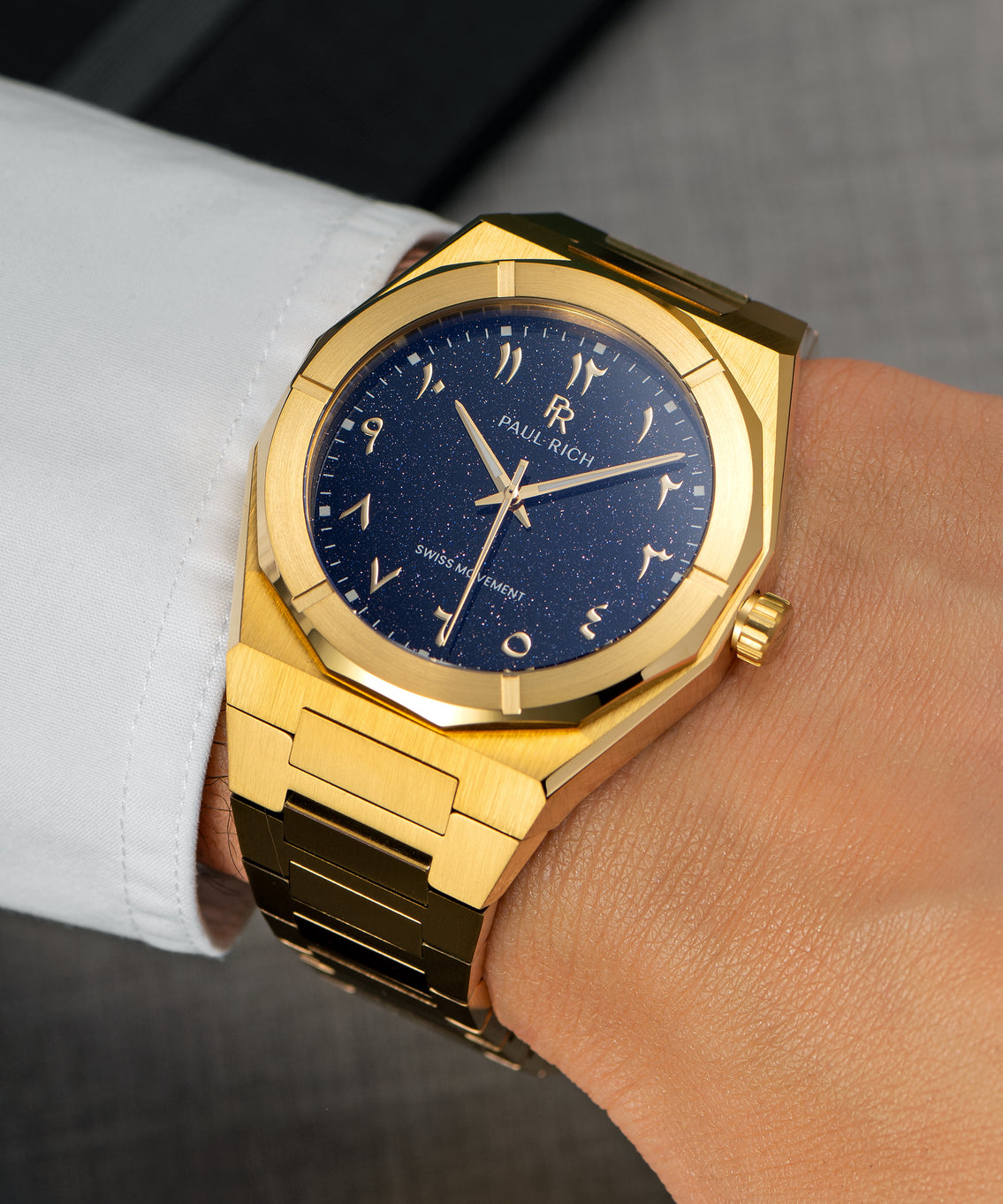 Paul Rich watch with brushed gold case and blue aventurine dial with Arabic numerals, worn with a white cuff over a grey suit. Paul Rich watch with brushed gold case and blue aventurine dial with Arabic numerals, worn with a white cuff over a grey suit.
