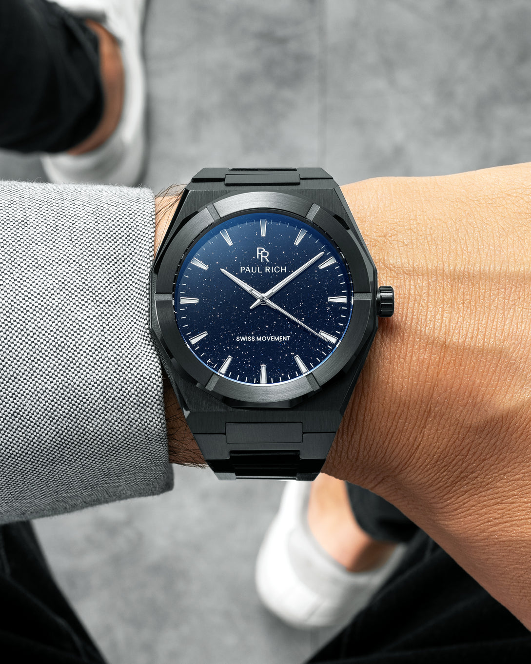 Black Paul Rich watch with blue aventurine dial worn on wrist, paired with light grey shirt and black pants, white sneakers visible. Black Paul Rich watch with blue aventurine dial worn on wrist, paired with light grey shirt and black pants, white sneakers visible.