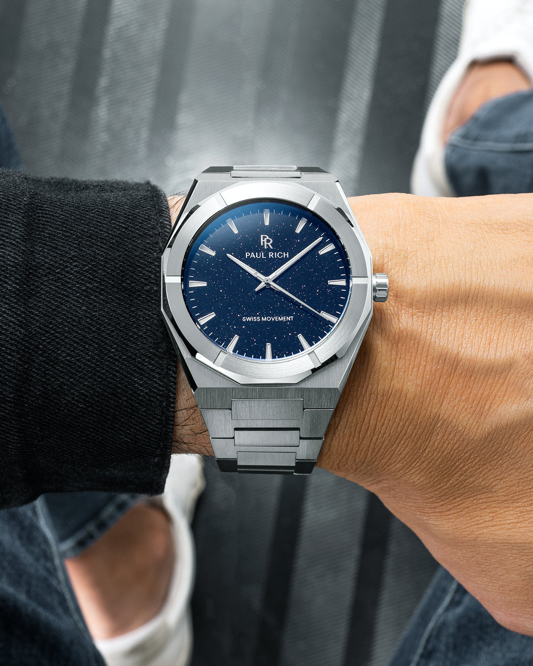Silver Paul Rich watch with blue aventurine dial on wrist, worn over black sleeve with jeans and white sneakers visible. Silver Paul Rich watch with blue aventurine dial on wrist, worn over black sleeve with jeans and white sneakers visible.