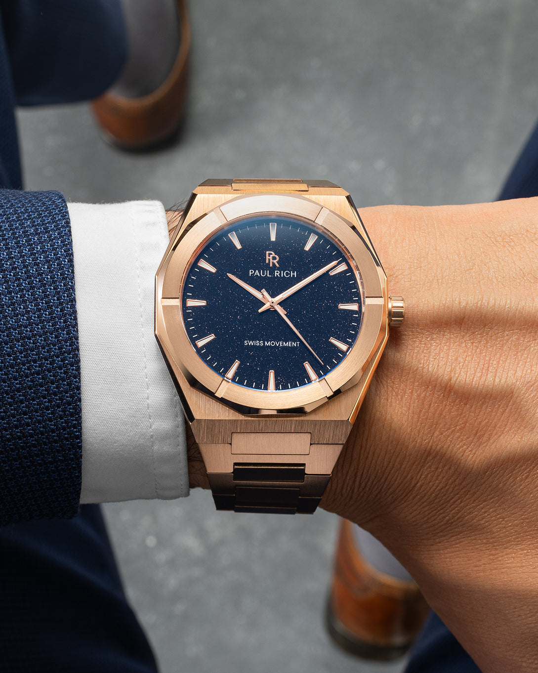 Rose gold Paul Rich watch with blue aventurine dial worn on wrist, paired with navy suit and white shirt, brown shoes in background. Rose gold Paul Rich watch with blue aventurine dial worn on wrist, paired with navy suit and white shirt, brown shoes in background.