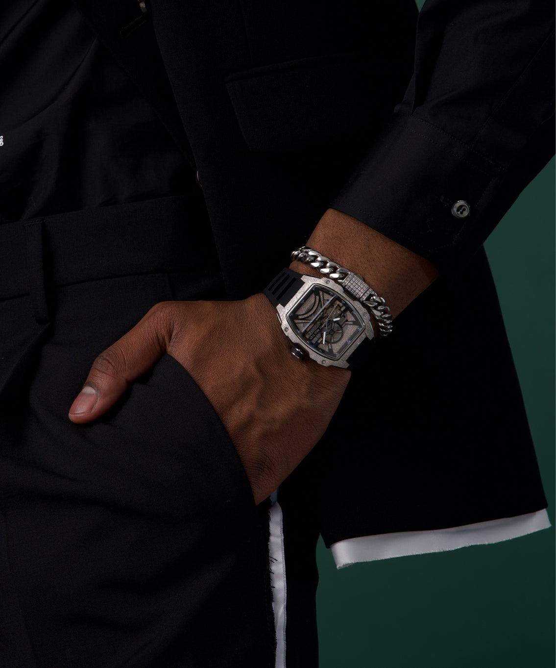 Man wearing a silver skeleton-dial Paul Rich watch with black strap and silver Cuban link bracelet, dressed in a black suit. Man wearing a silver skeleton-dial Paul Rich watch with black strap and silver Cuban link bracelet, dressed in a black suit.