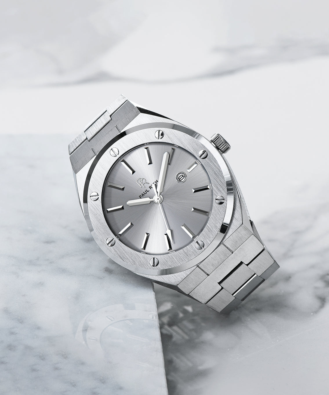 Paul Rich brushed silver watch with metallic sunray dial and date window, resting on white marble surface with soft reflections. Paul Rich brushed silver watch with metallic sunray dial and date window, resting on white marble surface with soft reflections.