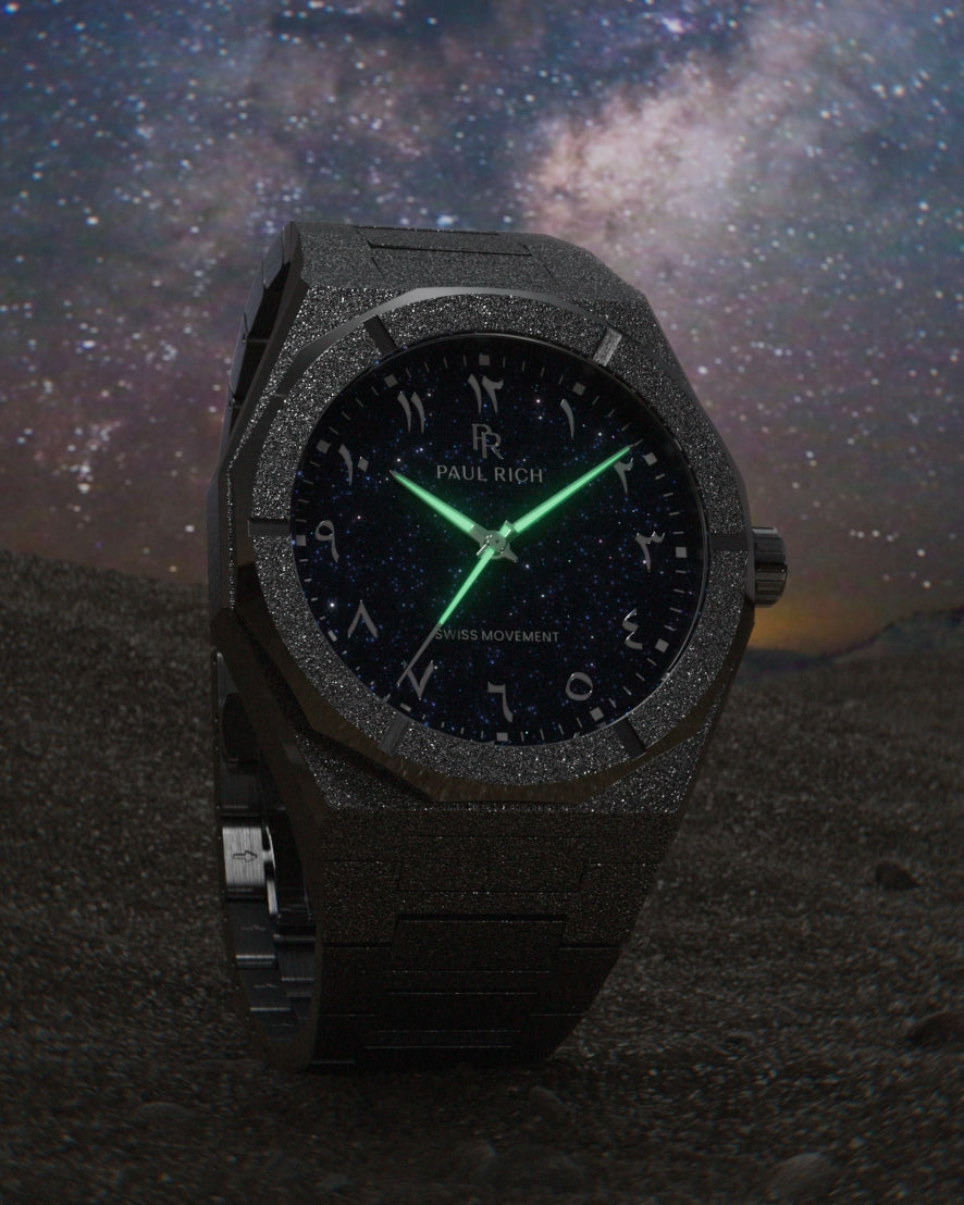 Frosted black watch with blue aventurine dial, Arabic numerals, and glowing green hands, set against a starry night sky on a dark terrain. Frosted black watch with blue aventurine dial, Arabic numerals, and glowing green hands, set against a starry night sky on a dark terrain.