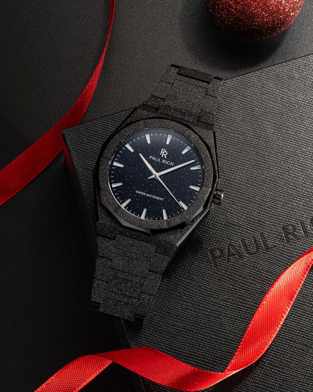 Paul Rich watch with frosted black case and black aventurine dial, displayed on a textured gift box with red ribbon and festive decor. Paul Rich watch with frosted black case and black aventurine dial, displayed on a textured gift box with red ribbon and festive decor.