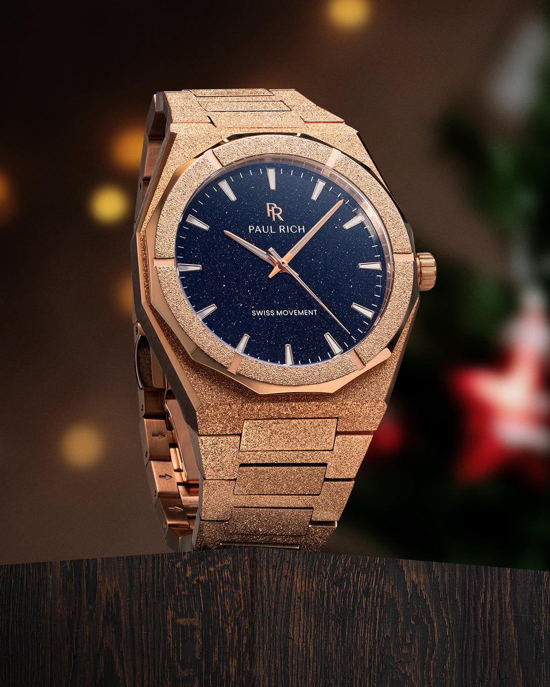 Rose gold Paul Rich watch with blue aventurine dial on wood surface, warm lights and blurred Christmas decor in the background. Rose gold Paul Rich watch with blue aventurine dial on wood surface, warm lights and blurred Christmas decor in the background.
