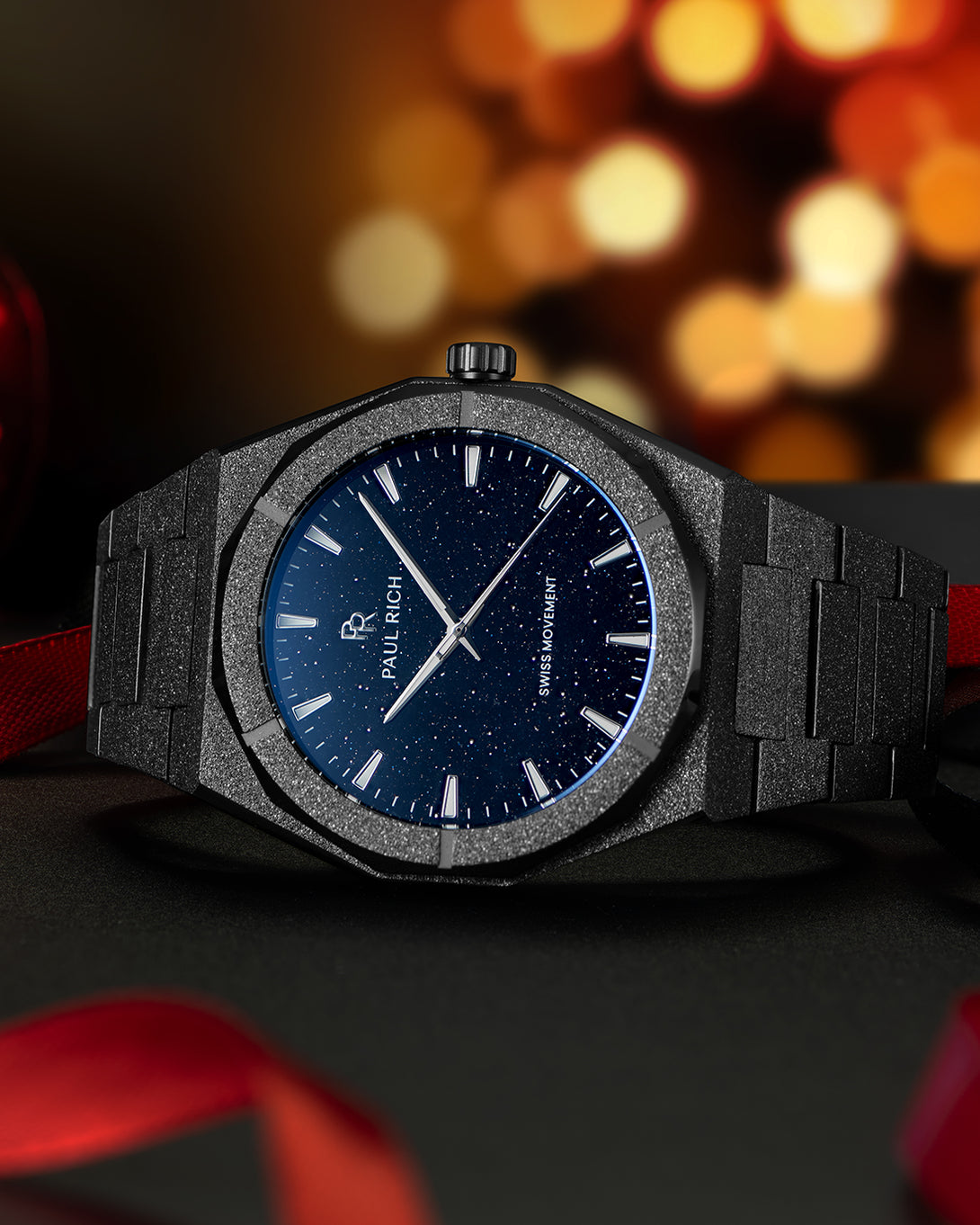 Black frosted Paul Rich watch with blue aventurine dial on black surface, surrounded by red ribbon and warm holiday lights in background. Black frosted Paul Rich watch with blue aventurine dial on black surface, surrounded by red ribbon and warm holiday lights in background.