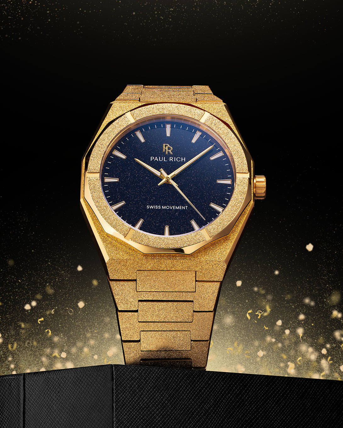Gold frosted Paul Rich watch with blue aventurine dial displayed upright on textured black surface, with golden sparkles rising behind. Gold frosted Paul Rich watch with blue aventurine dial displayed upright on textured black surface, with golden sparkles rising behind.