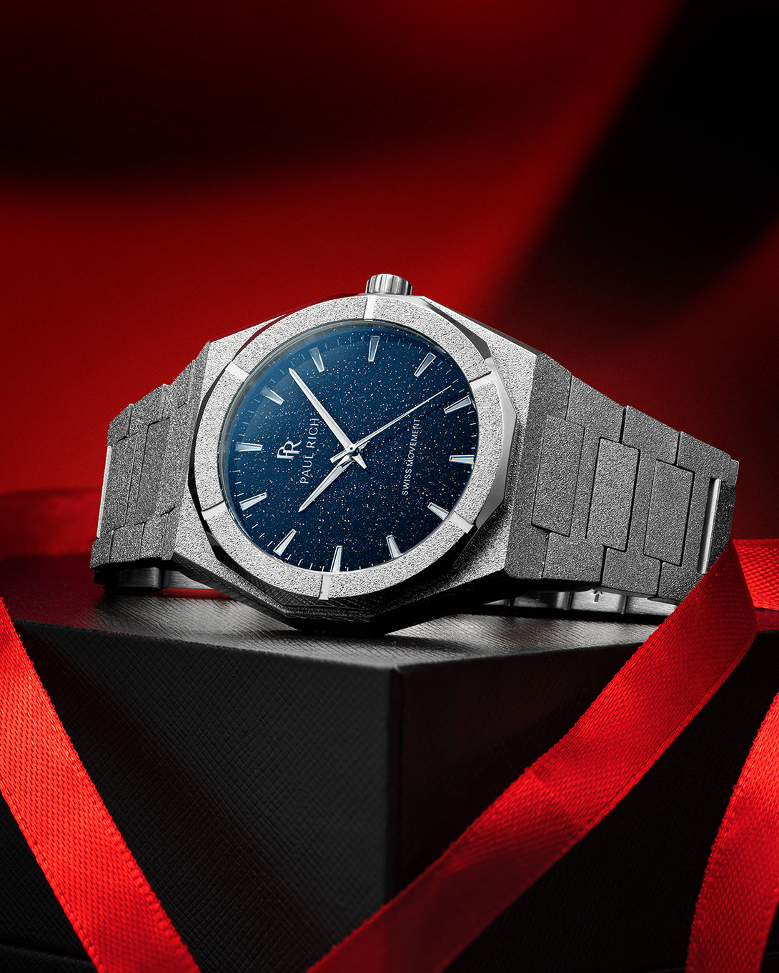 Silver frosted Paul Rich watch with a deep blue aventurine dial, placed on a black gift box with red ribbon on red-black background. Silver frosted Paul Rich watch with a deep blue aventurine dial, placed on a black gift box with red ribbon on red-black background.