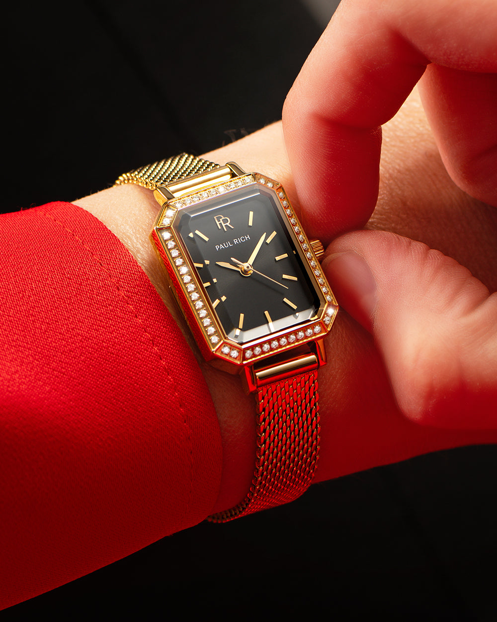 Gold-tone Paul Rich watch with black sunray dial, gem-set octagonal bezel, and gold mesh strap, worn on wrist with a red sleeve while a hand adjusts the crown. Gold-tone Paul Rich watch with black sunray dial, gem-set octagonal bezel, and gold mesh strap, worn on wrist with a red sleeve while a hand adjusts the crown.