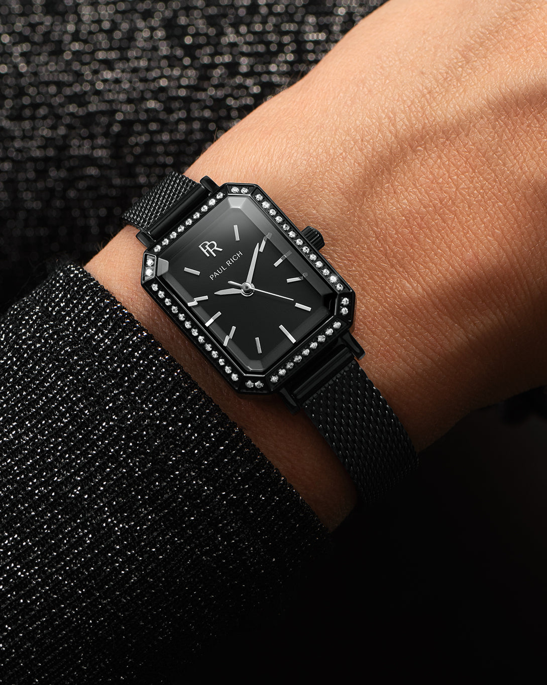 Black Paul Rich watch with black sunray dial, gem-set octagonal bezel, and black mesh strap, worn on wrist with a sparkling black sleeve. Black Paul Rich watch with black sunray dial, gem-set octagonal bezel, and black mesh strap, worn on wrist with a sparkling black sleeve.