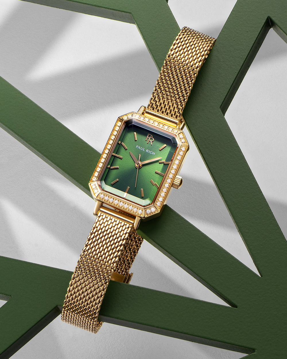 Gold-tone Paul Rich watch with a solid emerald green sunray dial, gem-set octagonal bezel, and gold mesh strap, displayed draped over a dark green geometric sculpture on a light background. Gold-tone Paul Rich watch with a solid emerald green sunray dial, gem-set octagonal bezel, and gold mesh strap, displayed draped over a dark green geometric sculpture on a light background.